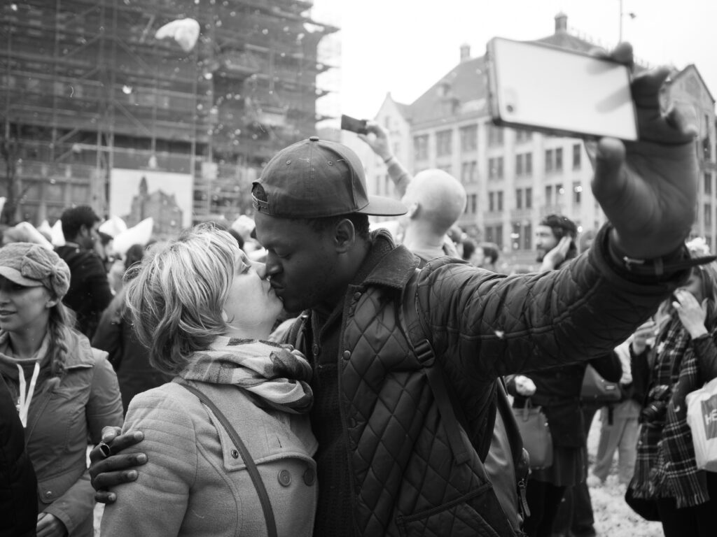 A couple in love making a kissing selfie in front of a massive pillow fight on Dam Square in Amsterdam on International Pillow Fight Day 2016.